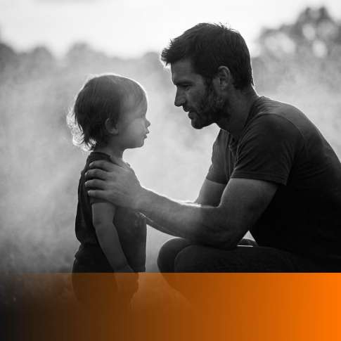 MenTools blog feature image for how to be a present dad when you work full time, black and white photograph of a father crouched at his young child's eye level in warm backlight, orange MenTools overlay band