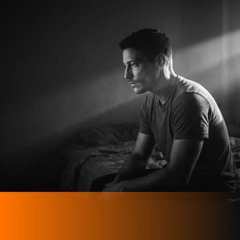 Tired man in his early thirties sitting on the edge of an unmade bed in a dark bedroom, MenTools blog feature image for the complete sleep guide for men