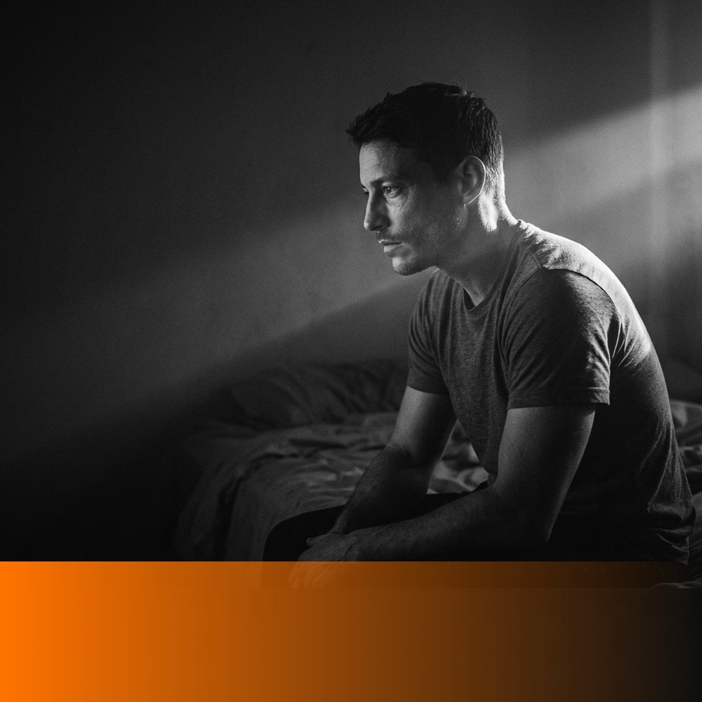 Tired man in his early thirties sitting on the edge of an unmade bed in a dark bedroom, MenTools blog feature image for the complete sleep guide for men
