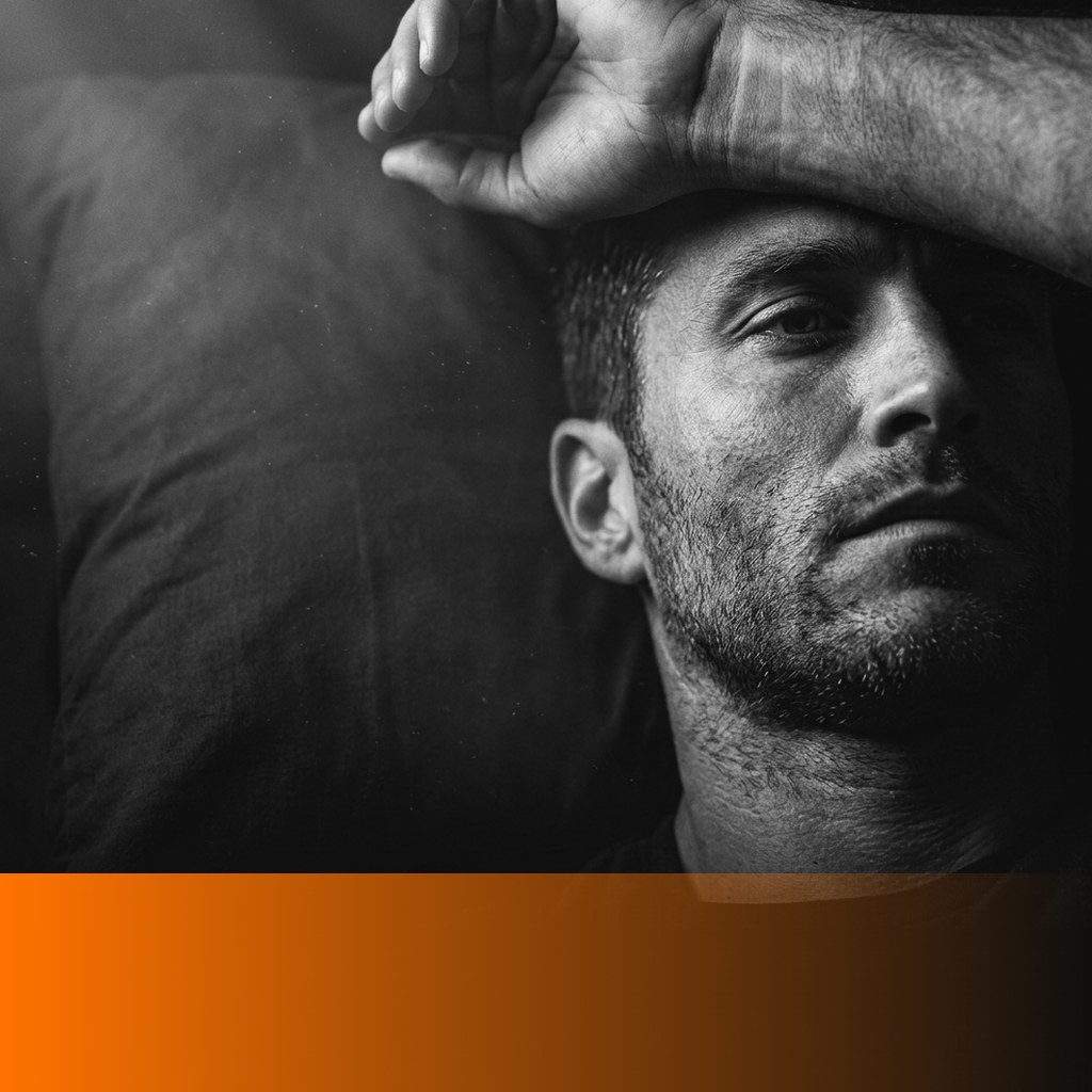 Man in his mid thirties lying awake in a dark bedroom with a forearm across his brow and his eyes open and unfocused, MenTools blog feature image for an article on why men feel wired but tired